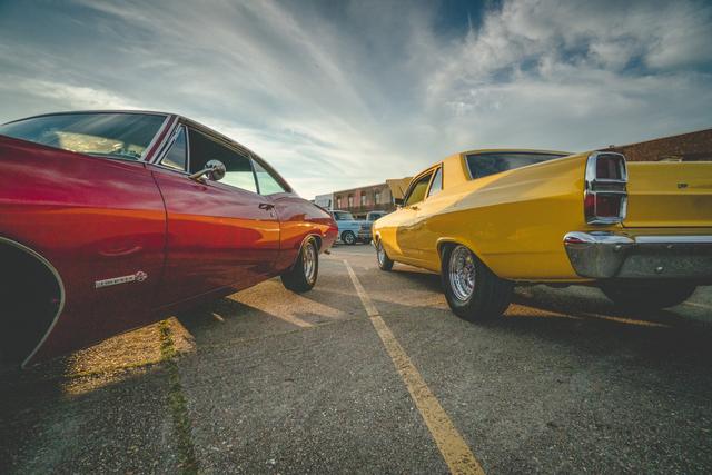 Low-angle wide shot of a red Chevy Impala beside a yellow Ford Fairlane at a car show under sweeping clouds